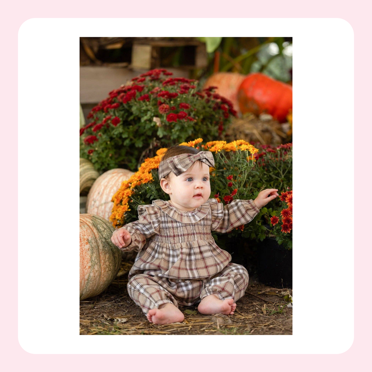 A baby wearing a brown and beige plaid smocked romper and matching headband sits outdoors among pumpkins and red and orange flowers.