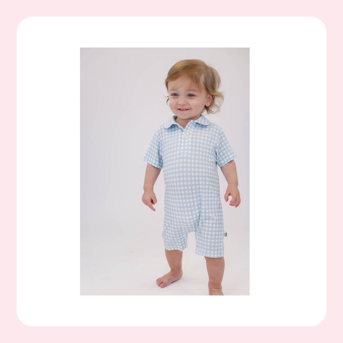 A toddler standing barefoot in a light blue and white gingham short-sleeve romper, centered within a white rounded square on a pale pink background.