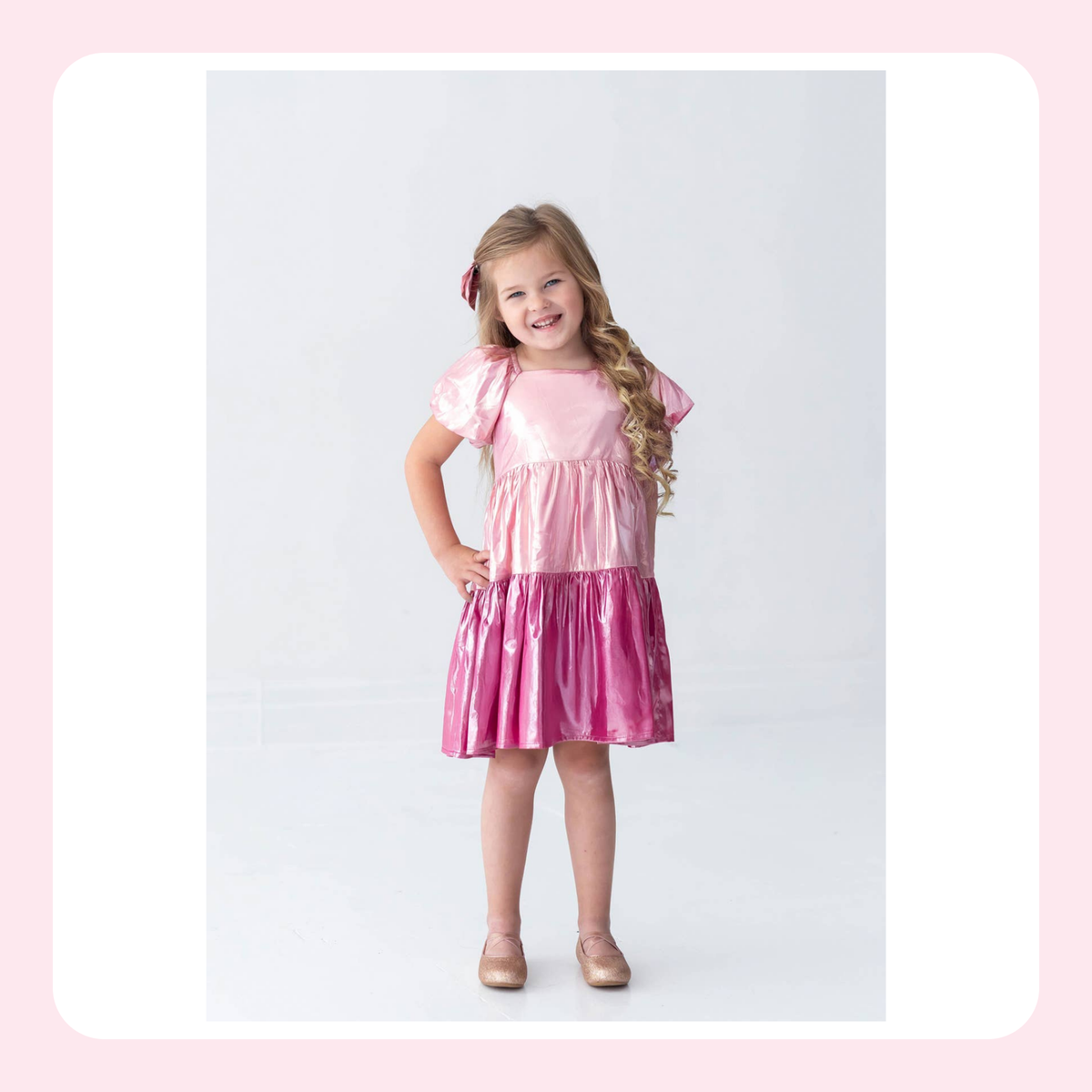 A young girl with long blonde hair wearing a shiny pink dress with puffed short sleeves, standing against a light gray studio background with a white and pink border.