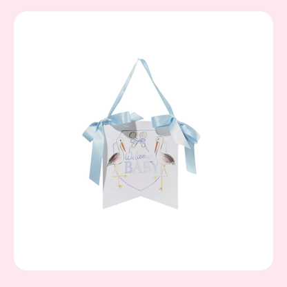 A white 'Welcome Baby' hanging banner with a light blue ribbon handle, decorated with two stork illustrations and the text 'We love BABY'.
