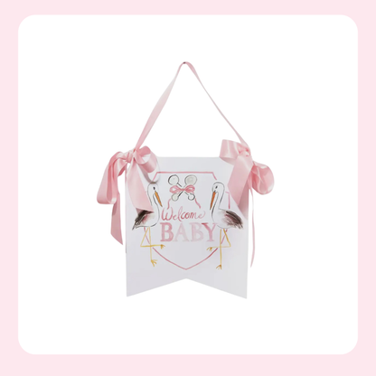 A white hanging banner sign with a pink ribbon hanger, featuring a pink and gold 'Welcome BABY' design with cartoon pelicans and bows.