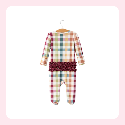 A baby long-sleeve footed one-piece jumpsuit with a multicolor plaid pattern and a dark burgundy ruffled waistband, hanging on a wooden hanger against a white background with a soft pink border.