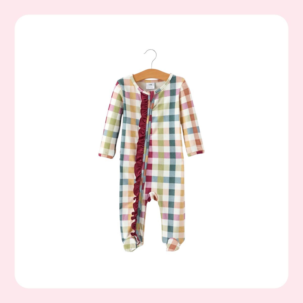 A baby footed sleeper with a pastel multicolor plaid pattern and a dark red ruffle trim down the front, hanging on a wooden hanger against a white background with a light pink border.