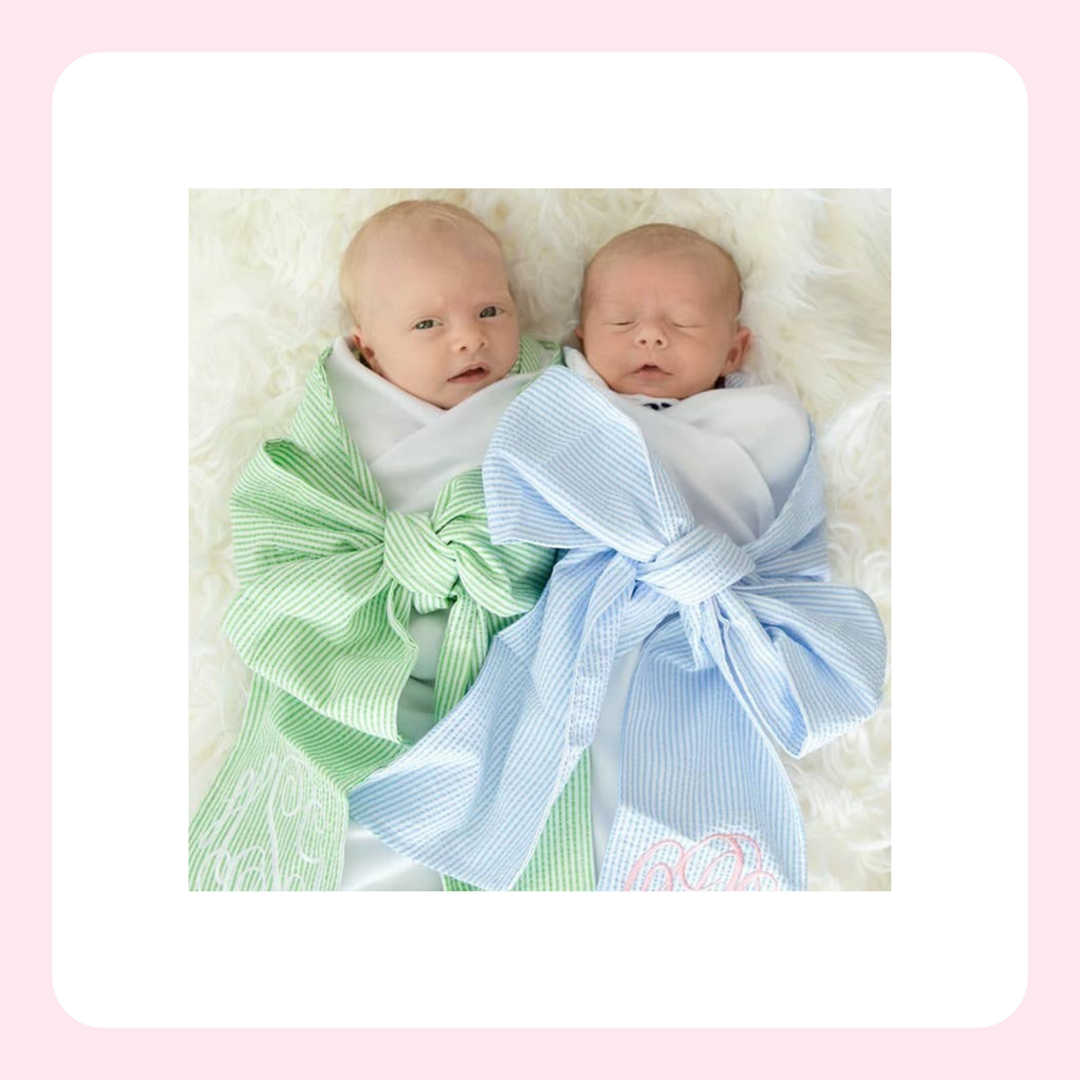 Two newborn babies lying side by side on a white faux fur surface, swaddled in large striped fabric bows in light green and light blue. The photo is centered within a white rounded square on a soft pink background.