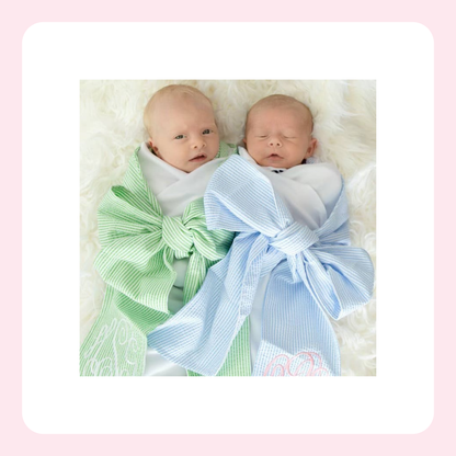 Two newborn babies lying side by side on a white faux fur surface, swaddled in large striped fabric bows in light green and light blue. The photo is centered within a white rounded square on a soft pink background.