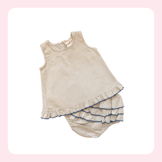 A two-piece baby outfit set in a light beige corduroy fabric, featuring a sleeveless top with a ruffled hem and matching ruffled bloomers with blue piping trim, displayed on a white background with a pink border.