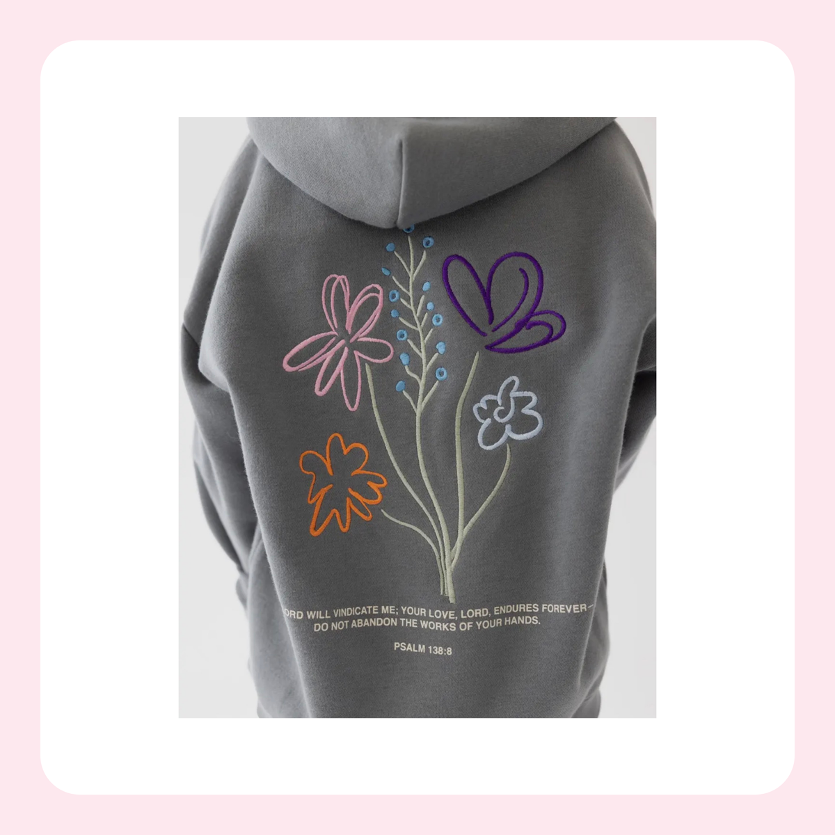 A dark gray hoodie shown from the back, featuring embroidered flowers and a butterfly in pink, purple, orange, and white, with a bible verse printed below.