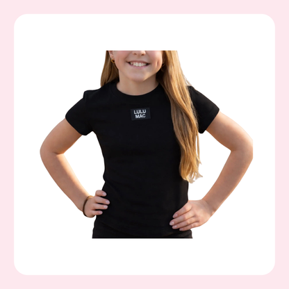 A young girl with long blonde hair wearing a black short-sleeve t-shirt with a small black square logo on the left chest that reads 'LULU MAC' in white text, posed with hands on hips against a white background with a light pink border.