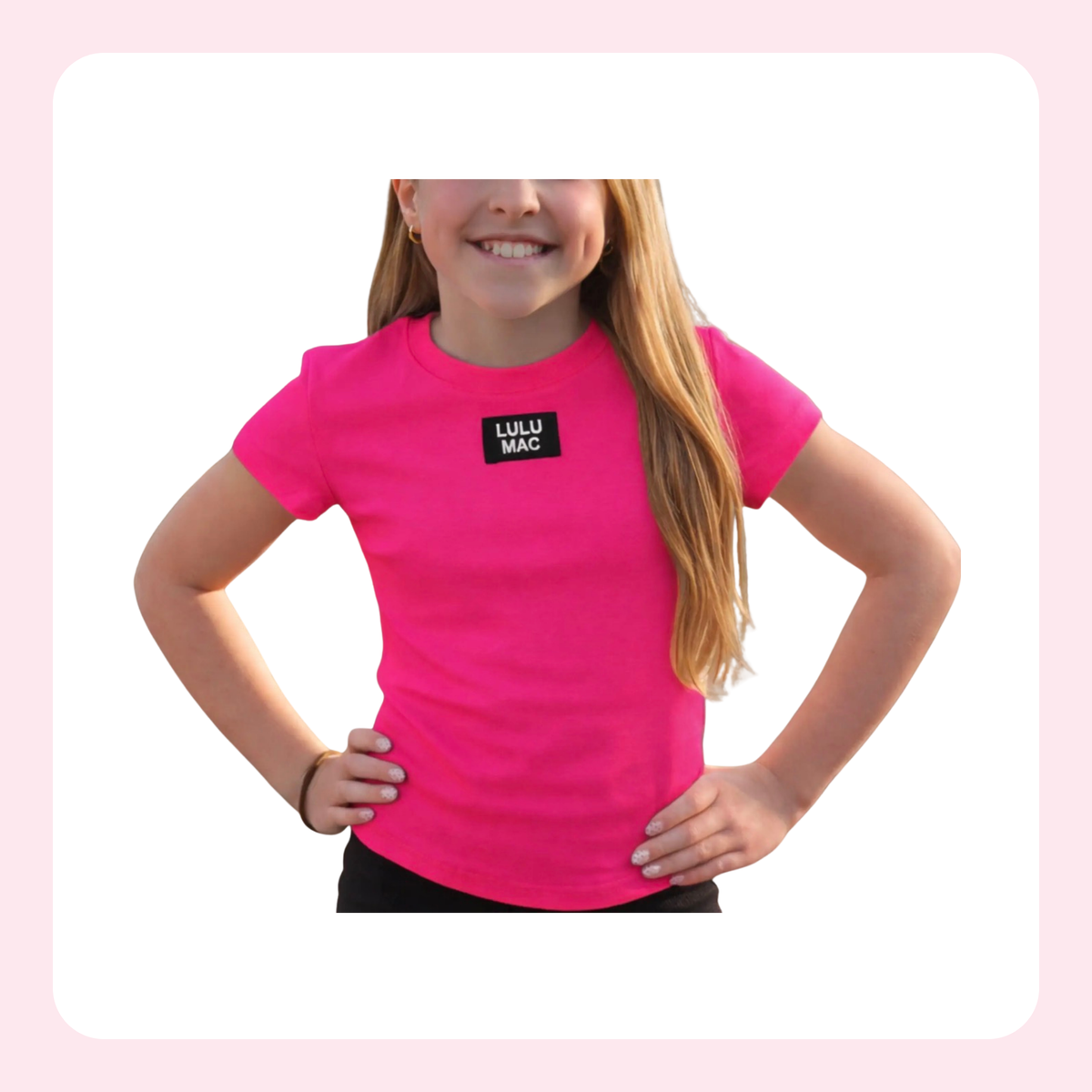 A young girl with long blonde hair wearing a bright pink short-sleeve t-shirt with a black square logo on the chest that reads 'LULU MAC'. She is posing with her hands on her hips against a white background with a light pink border.