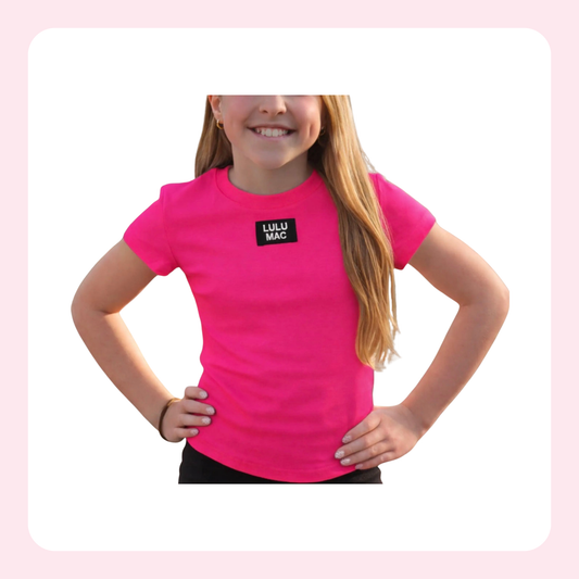 A young girl with long blonde hair wearing a bright pink short-sleeve t-shirt with a black square logo on the chest that reads 'LULU MAC'. She is posing with her hands on her hips against a white background with a light pink border.