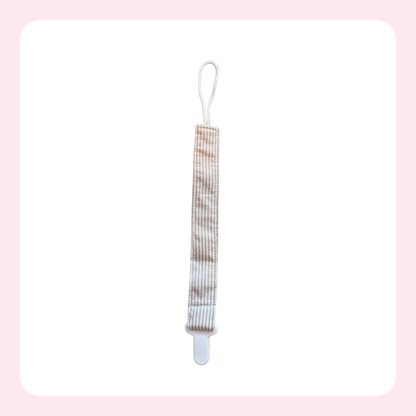 A baby pacifier clip with a beige and white striped fabric strap, white plastic clip, and white loop at the top, shown on a white background with a light pink border.