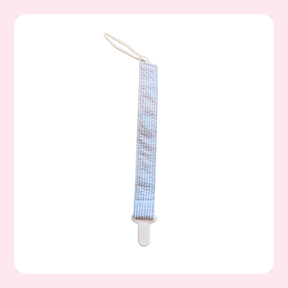 A baby pacifier clip with a light blue and white checkered patterned fabric strap and a white plastic clip, shown on a white background with a soft pink border.