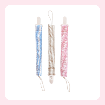 Three fabric baby pacifier clips with plastic snap hooks are shown vertically on a white background with a soft pink border. The left clip is light blue and white checkered, the middle is beige and white striped, and the right is pink and white checkered.