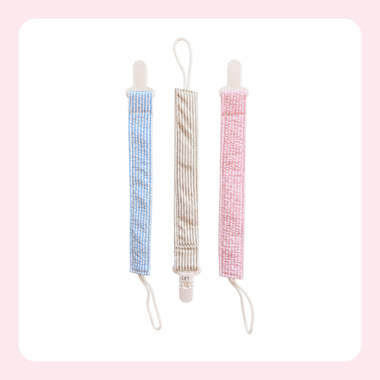 Three fabric baby pacifier clips with plastic snap hooks are shown vertically on a white background with a soft pink border. The left clip is light blue and white checkered, the middle is beige and white striped, and the right is pink and white checkered.