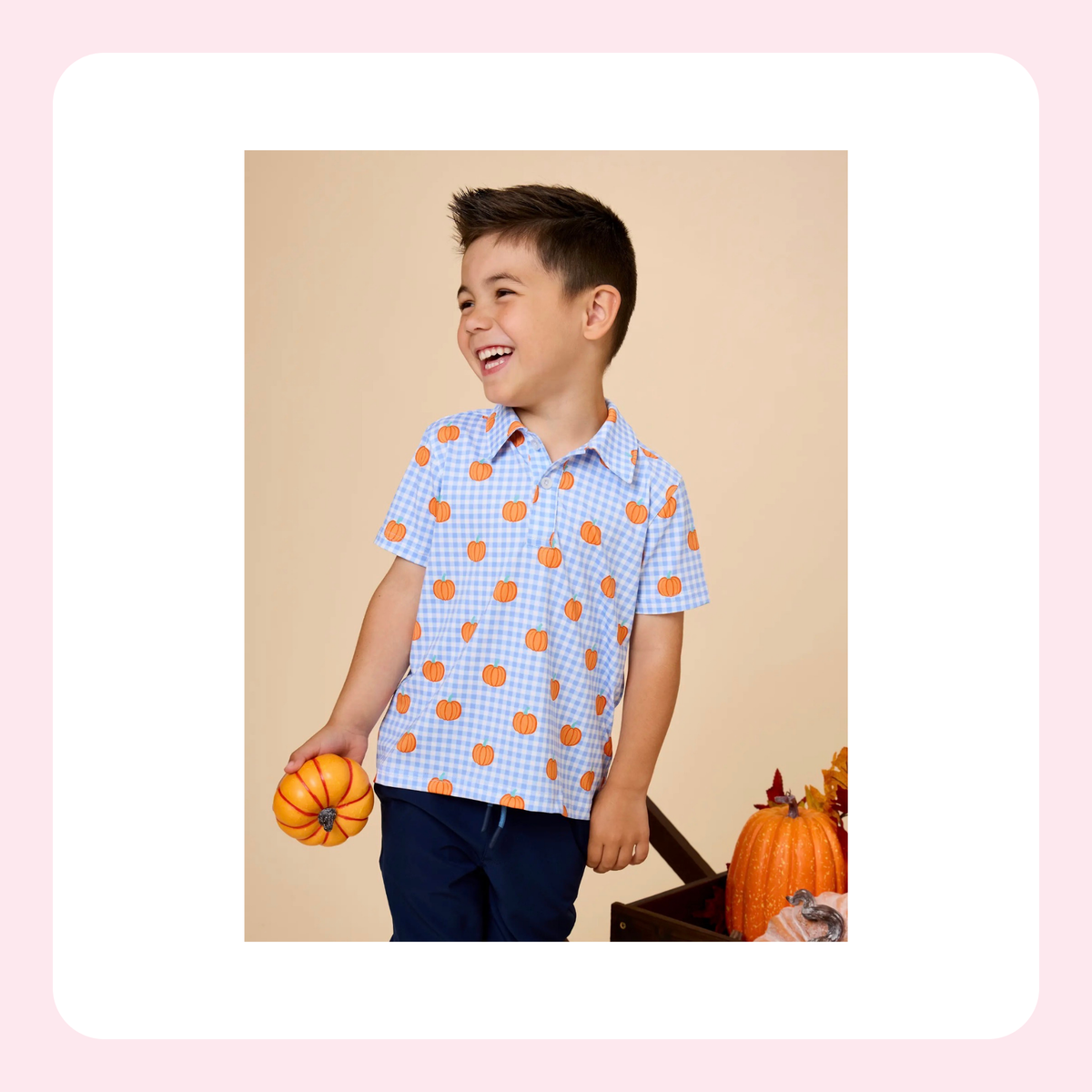 A young boy smiling and looking to the side, wearing a light blue and white gingham short-sleeve button-up shirt with orange pumpkin prints, paired with navy pants. He is holding a small orange pumpkin against a beige studio background with a decorative autumn basket.