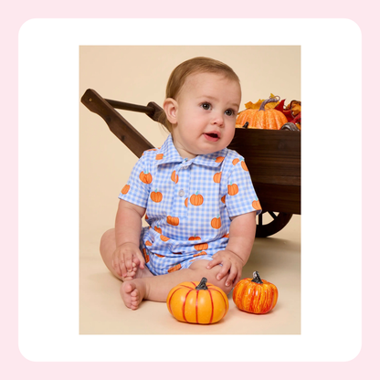 A baby wearing a light blue and white gingham short-sleeve romper with an orange pumpkin print, sitting on a beige floor next to two small decorative pumpkins in front of a rustic wooden wheelbarrow filled with autumn decorations.