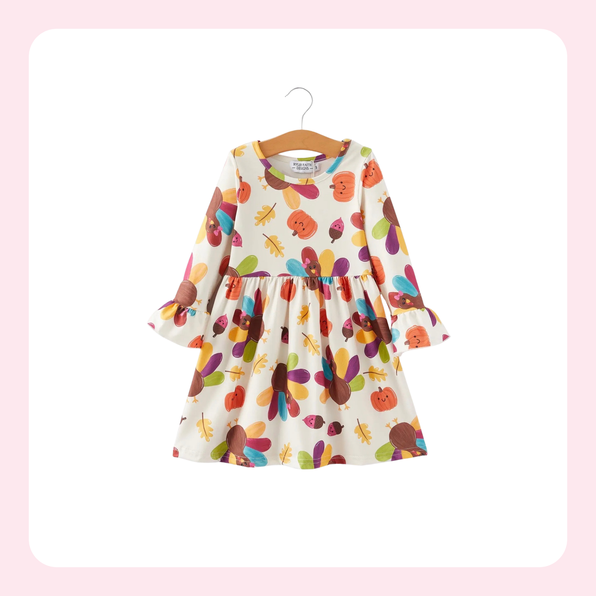 A long-sleeve baby dress with a colorful autumn leaf and pumpkin print on a cream background, hanging on a wooden hanger against a white background with a soft pink border.