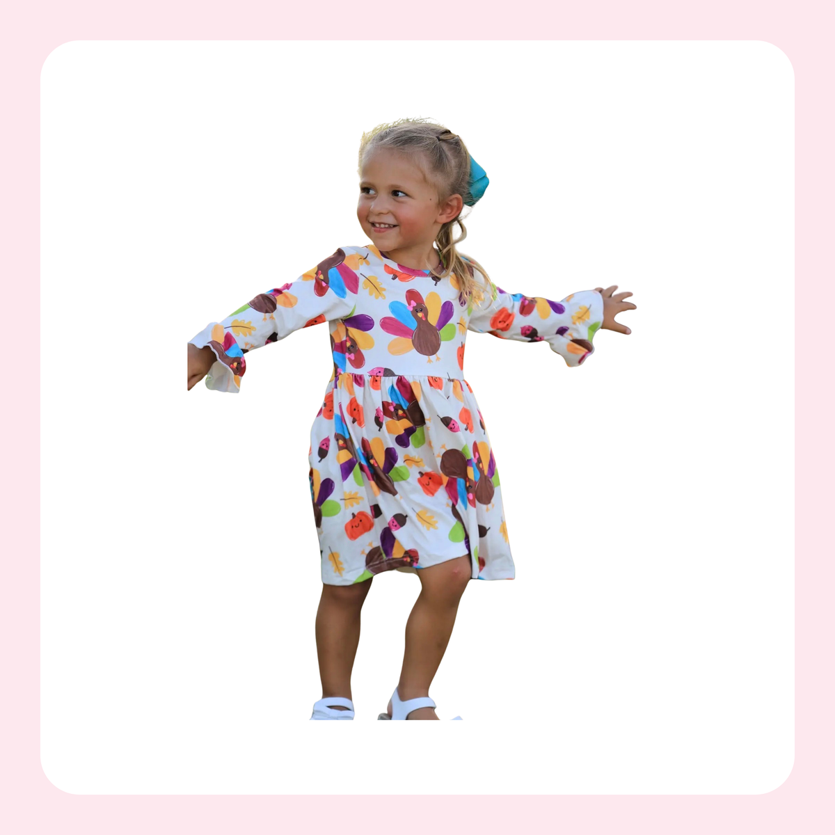 A young girl with blonde hair in a ponytail wearing a white long-sleeve dress with a colorful abstract pattern, standing with arms outstretched against a white background with a light pink border.