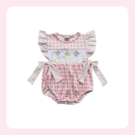 A baby girl's two-piece outfit featuring a pink and white gingham romper with ruffle sleeves and a matching long-sleeve top with a birthday balloon and cake graphic, displayed on a white background with a soft pink border.
