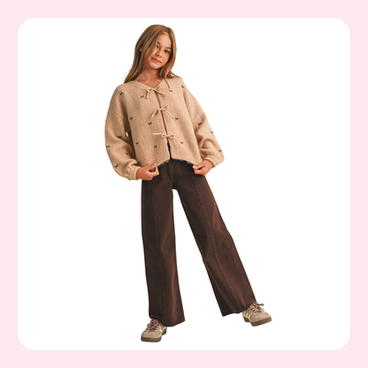 A young woman modeling a beige cropped knit cardigan with toggle closures and small embroidered details, paired with dark brown wide-leg pants and beige sneakers, set against a white background with a light pink border.