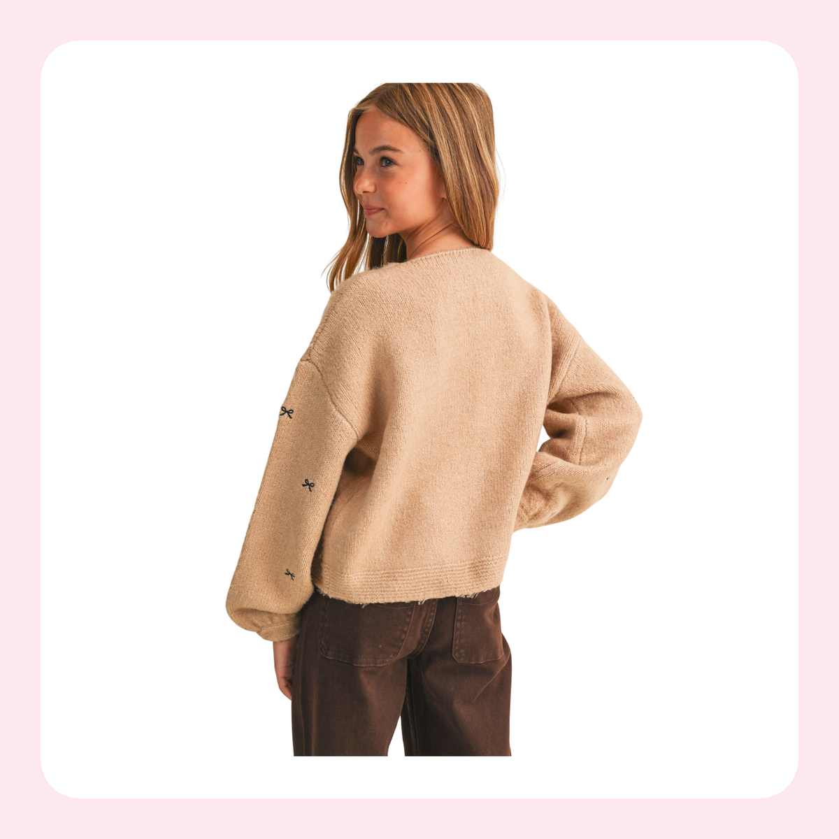 A young girl with blonde hair is shown from the back wearing a beige fuzzy cropped sweater with small embroidered details on the sleeves, paired with dark brown pants, set against a white background with a light pink border.