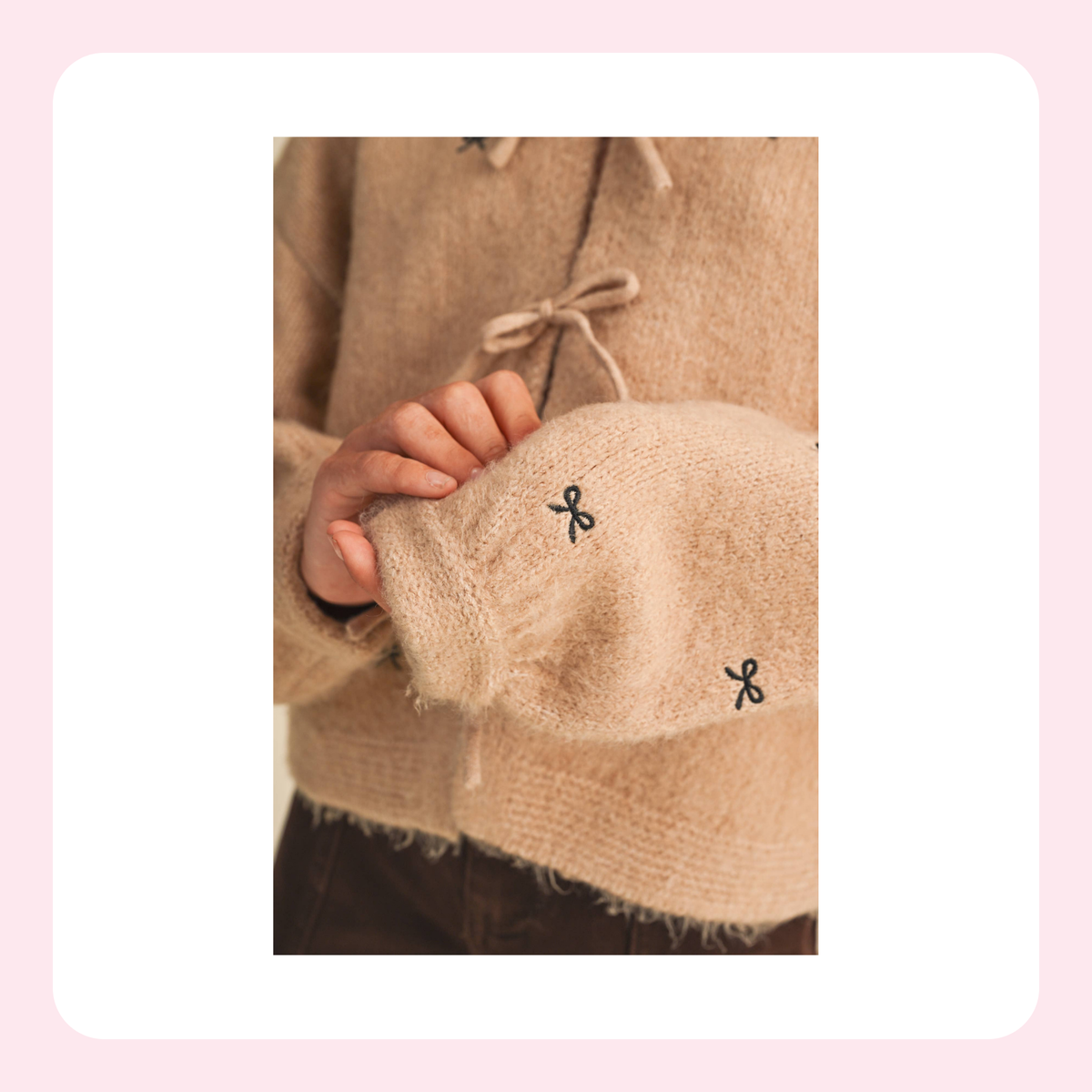 A close-up of a person wearing a fuzzy beige knit sweater with drawstring ties and small black bow embroidery on the sleeve, framed by a soft pink border.