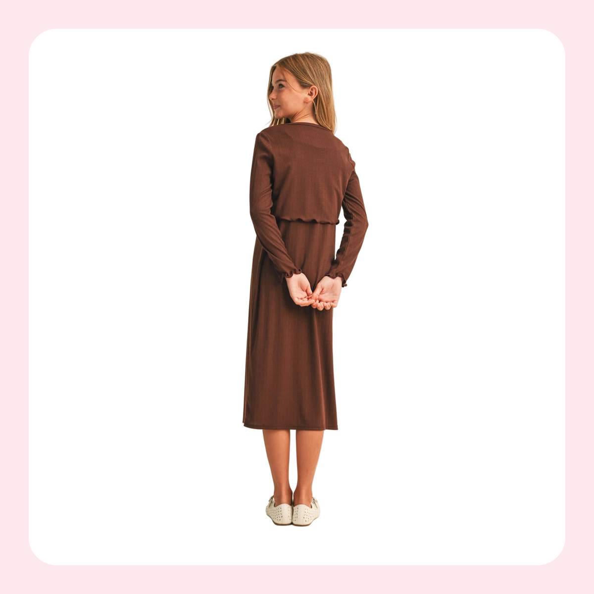 A young girl with blonde hair is shown from the back wearing a brown long-sleeve midi dress with a ruffled waist detail, paired with white slip-on shoes, set against a white background with a light pink border.