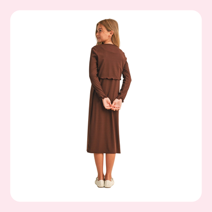 A young girl with blonde hair is shown from the back wearing a brown long-sleeve midi dress with a ruffled waist detail, paired with white slip-on shoes, set against a white background with a light pink border.