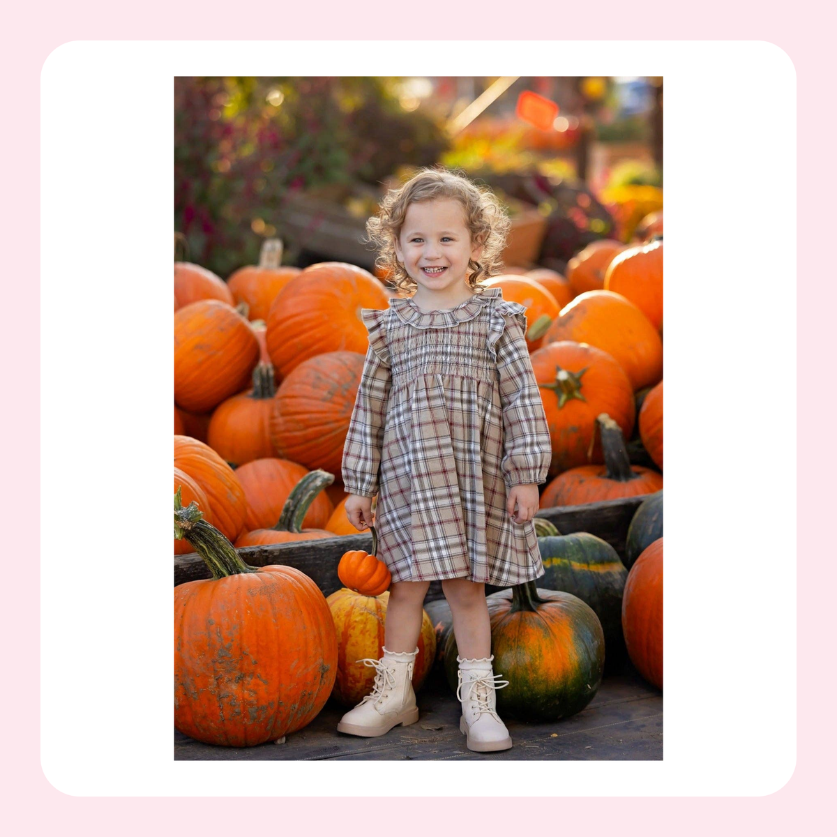 A young toddler girl stands in front of a large pile of pumpkins at a fall harvest display, wearing a beige and brown plaid smocked dress and cream lace-up boots.