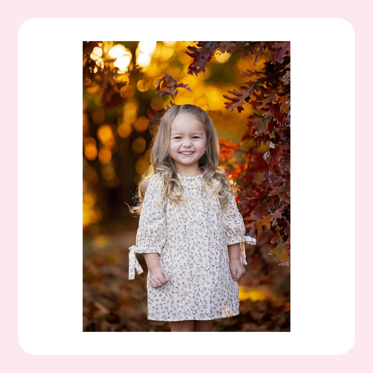 A young girl with long blonde hair stands outdoors in an autumn setting, wearing a cream-colored dress with a small floral pattern and tie details on the sleeves. The background features warm golden and red leaves.