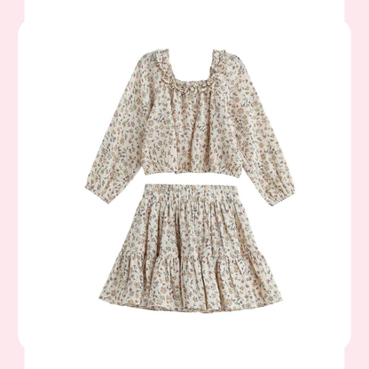 A two-piece girls' outfit set featuring a long-sleeve cropped blouse and a matching tiered mini skirt, both in a beige base fabric with a small multicolor floral print, shown on a white background with a decorative pink border.