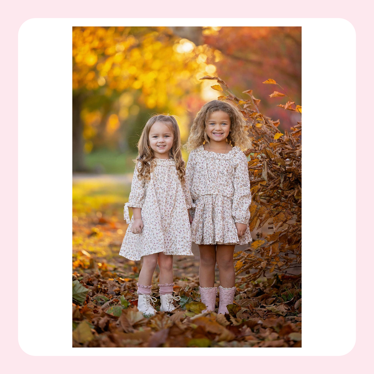 Two young girls standing outdoors in an autumn park, wearing matching long-sleeve floral dresses and pink boots, surrounded by fallen leaves and warm golden foliage.