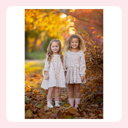 Two young girls standing outdoors in an autumn park, wearing matching long-sleeve floral dresses and pink boots, surrounded by fallen leaves and warm golden foliage.