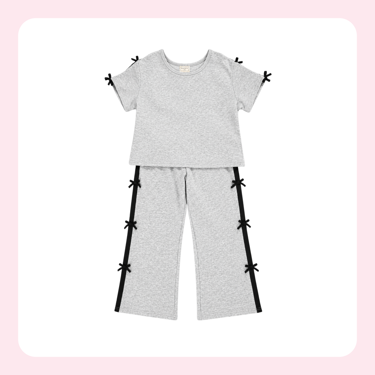A two-piece children's loungewear set in light heather gray, featuring a short-sleeve top with small black bow details at the sleeve cuffs and matching wide-leg pants with black side panels and bow accents, displayed on a white background with a soft pink border.