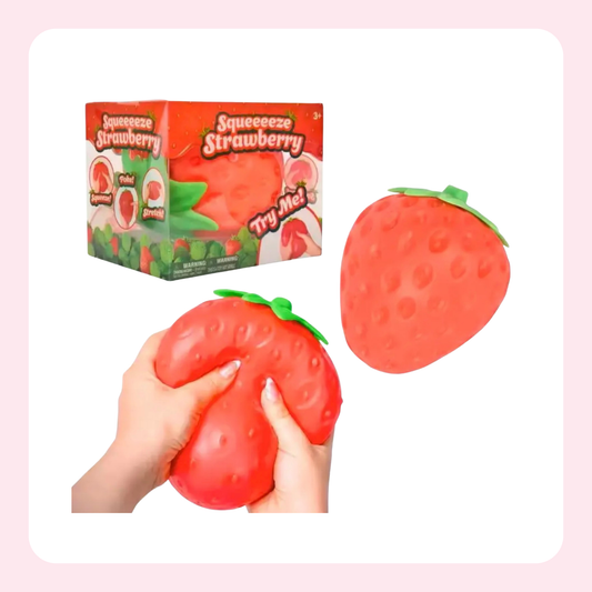A strawberry-shaped squeeze toy shown with its retail box and being held by two hands. The product is bright red with a green leafy top and textured surface, set against a white background with a light pink border.
