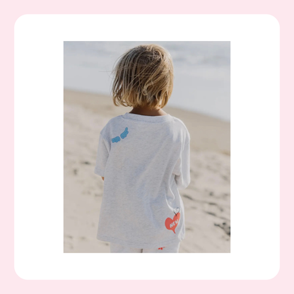A child viewed from behind wearing a light gray heather t-shirt with a blue butterfly graphic on the upper back and a red heart graphic on the lower back, standing on a sandy beach.