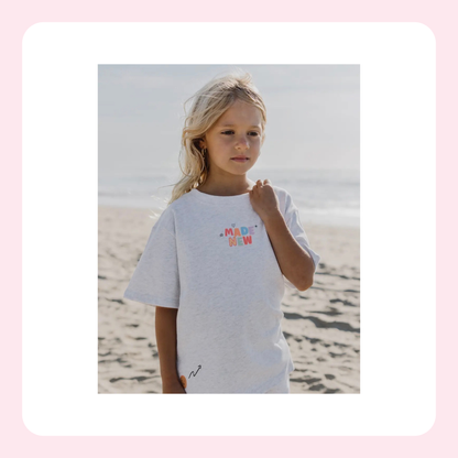 A young blonde girl wearing a white t-shirt with a colorful graphic on the chest, standing on a sandy beach with the ocean in the background.