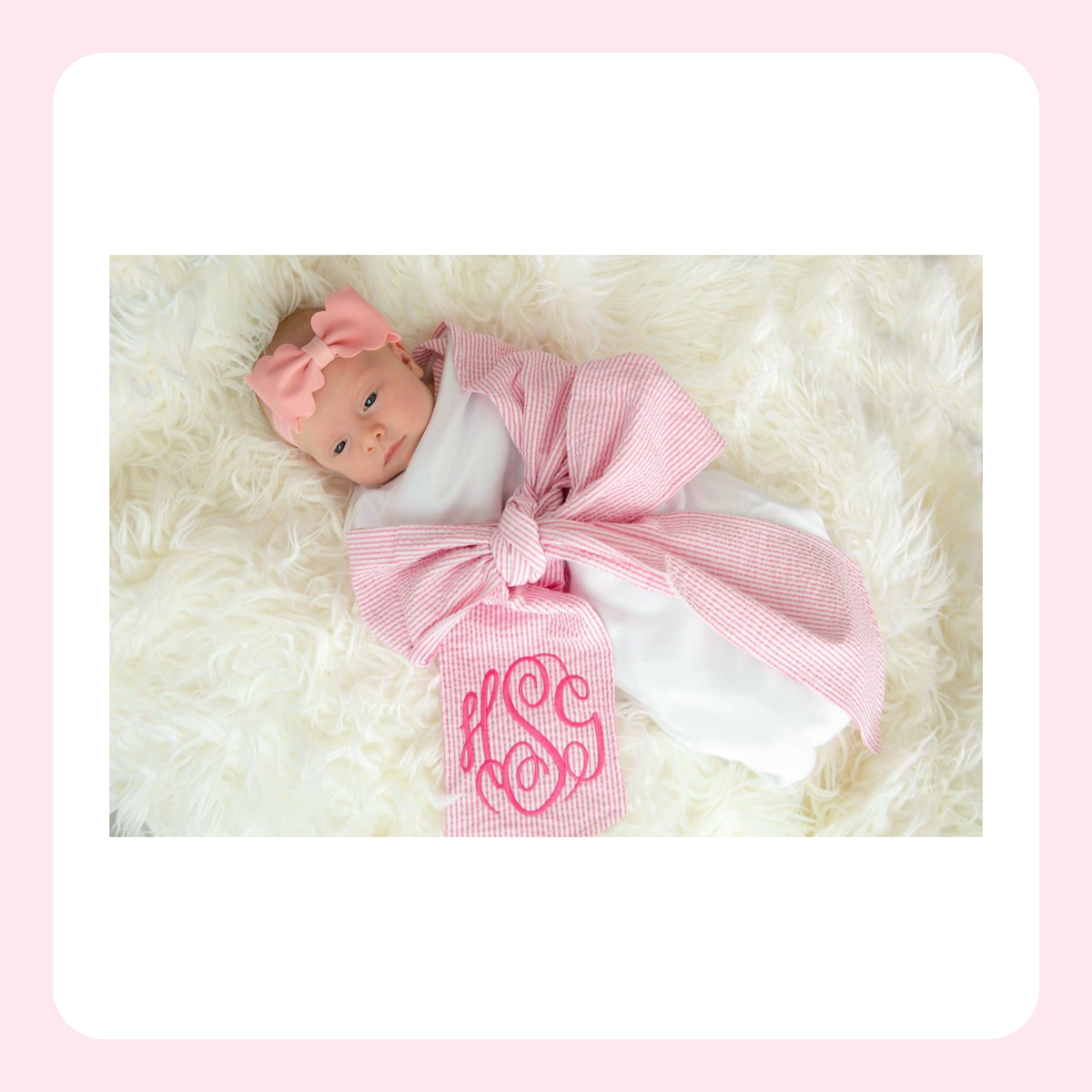 A baby doll wrapped in a pink and white gingham blanket with a large matching bow and monogram, lying on a white faux fur surface.