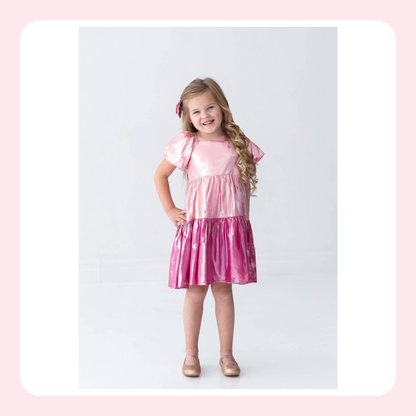 A young girl with long blonde hair wearing a shiny pink dress with puffed short sleeves, standing against a light gray studio background with a white and pink border.