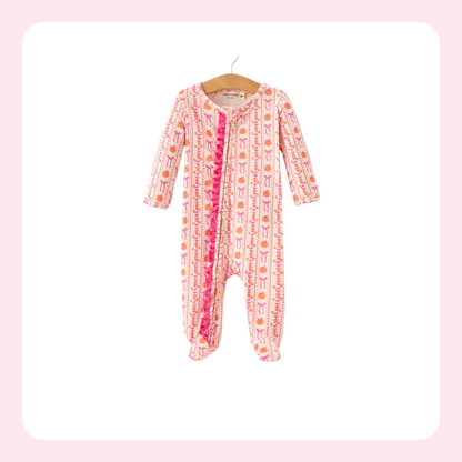 A baby footed sleeper with long sleeves and legs, featuring a pink and white patterned print with orange accents and a bright pink ruffle trim down the front, hanging on a wooden hanger against a white background with a light pink border.