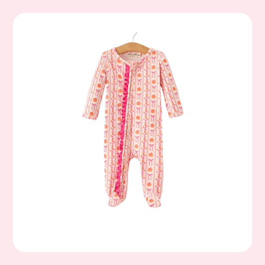 A baby footed sleeper with long sleeves and legs, featuring a pink and white patterned print with orange accents and a bright pink ruffle trim down the front, hanging on a wooden hanger against a white background with a light pink border.