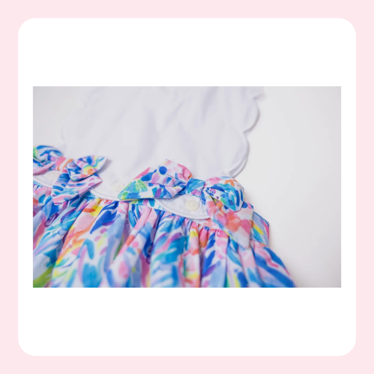 A close-up of a colorful children's dress with a white scalloped yoke and ruffled trim, shown against a soft white background with a light pink border.