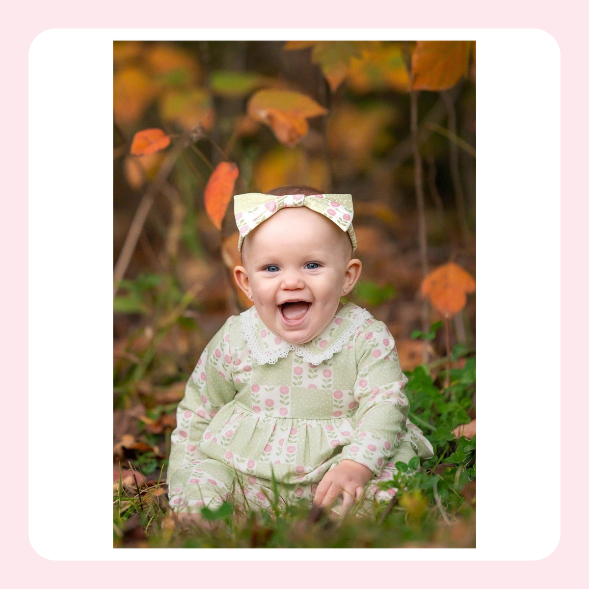 A baby wearing a light green and pink floral patterned dress with a matching bow headband, sitting outdoors in a grassy area with autumn leaves.