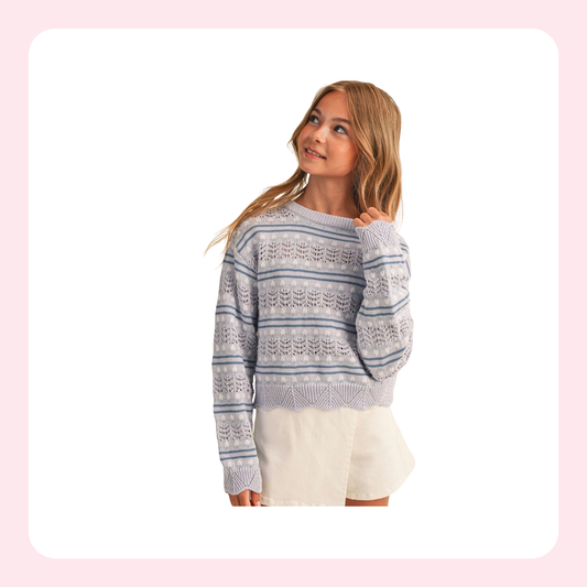 A young woman modeling a light blue and white patterned knit sweater with cream shorts against a white background with a soft pink border.