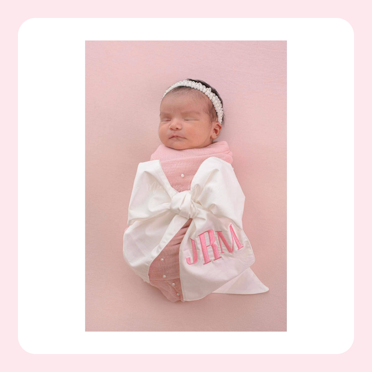 A newborn baby wrapped in a white blanket with a large bow and pink embroidered initials 'JHM', wearing a pink knit top and a white beaded headband, posed against a soft pink background.
