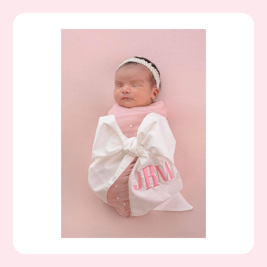 A newborn baby wrapped in a white blanket with a large bow and pink embroidered initials 'JHM', wearing a pink knit top and a white beaded headband, posed against a soft pink background.