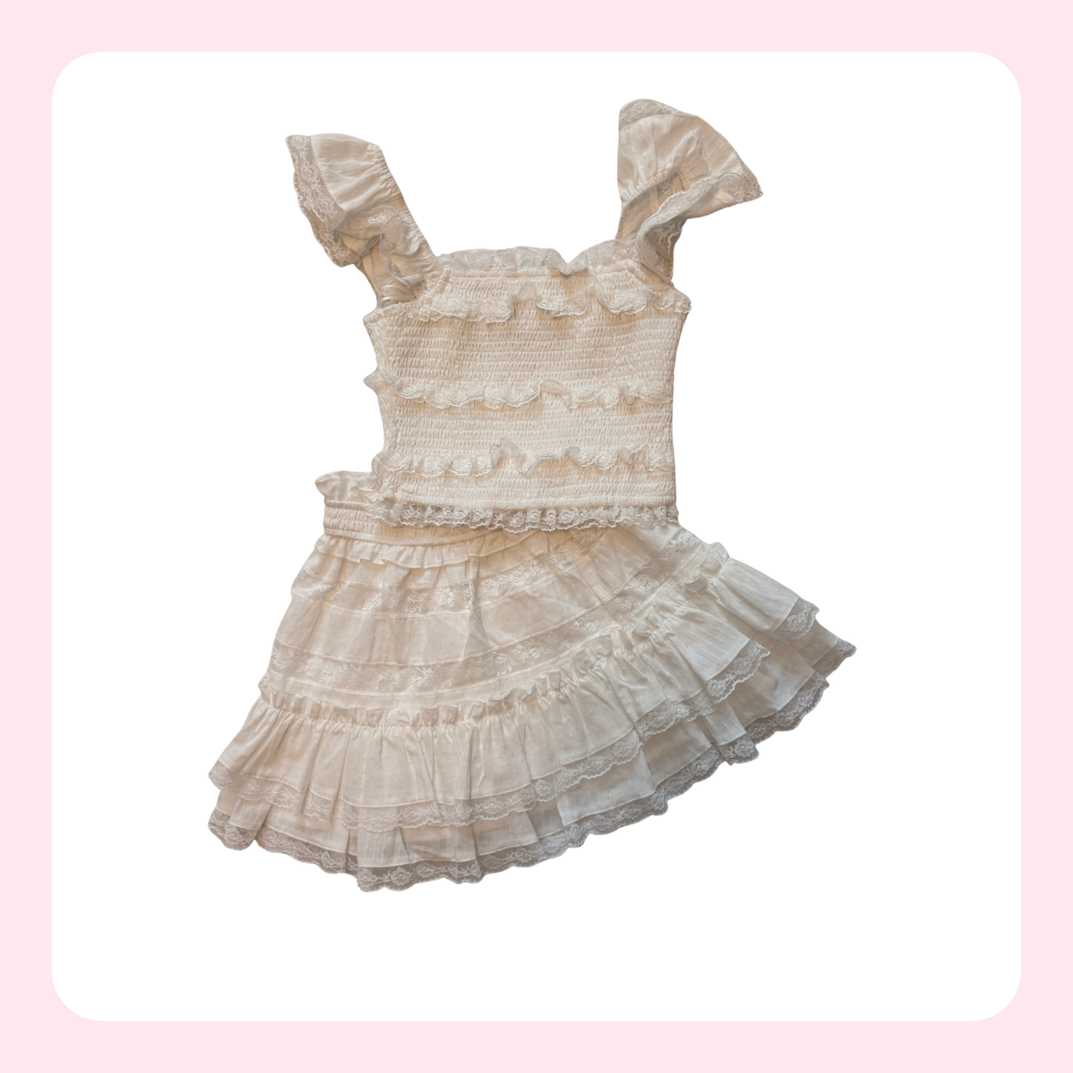 A cream-colored babydoll-style dress with ruffled details, smocked bodice, and tiered skirt, shown against a white background with a light pink border.
