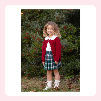 A young girl with curly blonde hair stands outdoors in front of dense green bushes with red berries, wearing a dark red cardigan over a white top, a green and navy plaid skirt, and white lace-up boots.