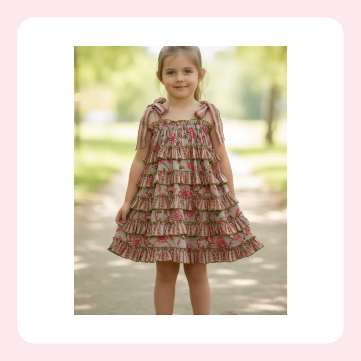 A young girl wearing a tiered ruffle dress with a pink and green floral print, standing outdoors on a blurred path.