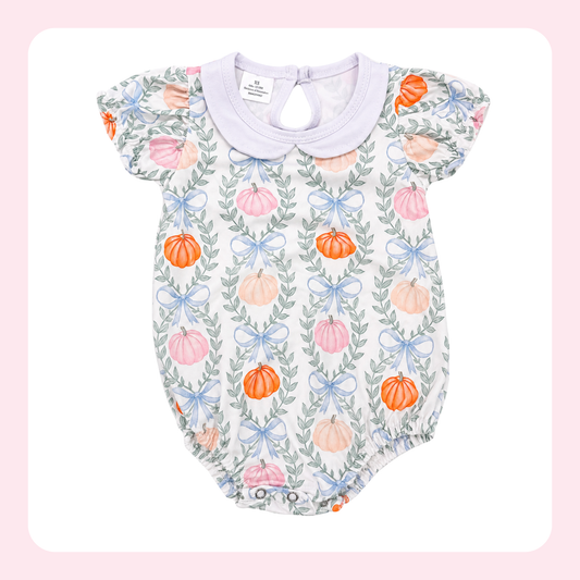 A baby romper with a white base, featuring a repeating pattern of orange and pink pumpkins, green leaves, and light blue bows, finished with a light purple collar.